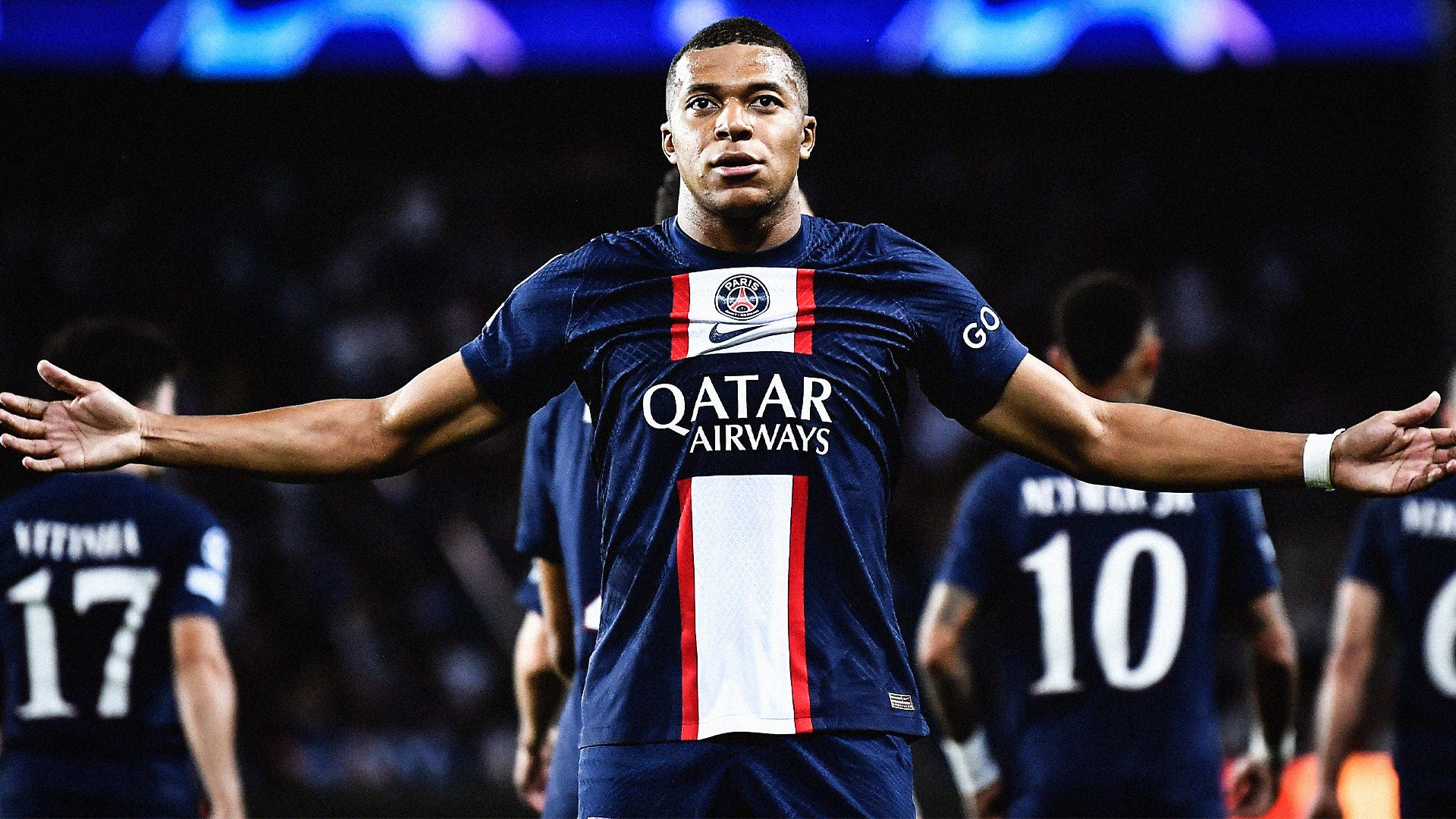  Kylian Mbappe is playing soccer for Paris Saint-Germain, wearing a blue and red jersey with white stripes, and celebrating a goal with his arms outstretched.