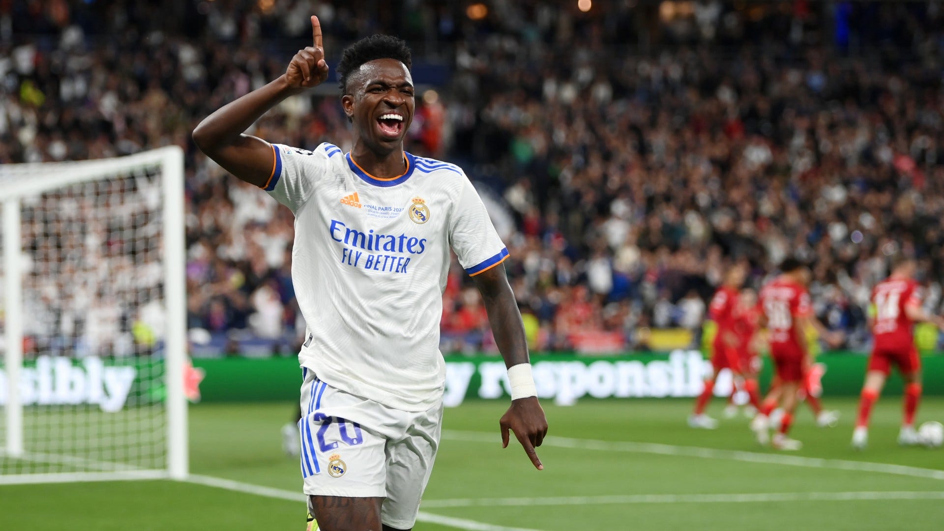  Vinicius Junior celebrates a goal for Real Madrid in the Champions League wearing a white jersey with blue trim and white shorts, surrounded by opposing players in red jerseys.